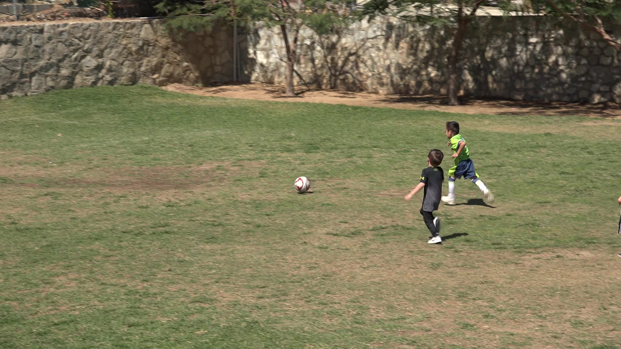 The boy in green and a child in black move farther across the grass in San Jose Del Cabo, Mexico, with the soccer ball rolling ahead near the center of the frame.