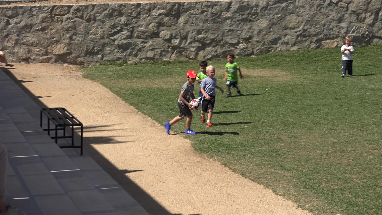 Small children and an adult stand near a soccer ball on a grass field in San Jose Del Cabo, Mexico, with a stone wall behind them.