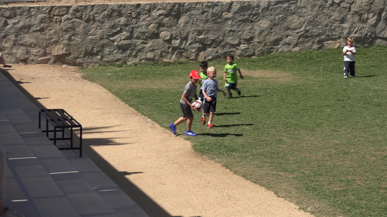 Children in casual sports clothes stand and walk near an adult coach and a soccer ball on the grass field in San Jose Del Cabo, Mexico.