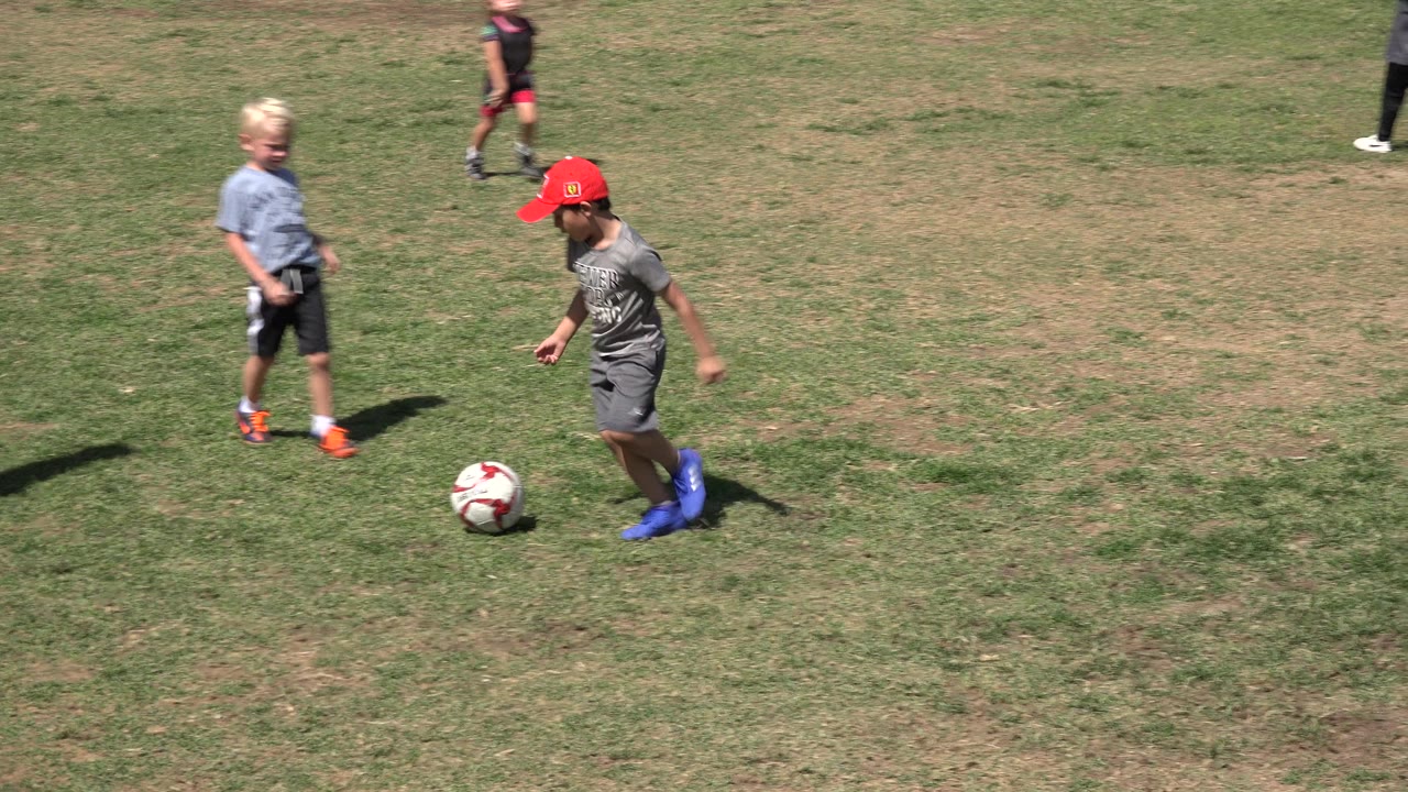 A child in a red cap steps toward a soccer ball on the grass in San Jose del Cabo, Mexico, as a blond child stands nearby.