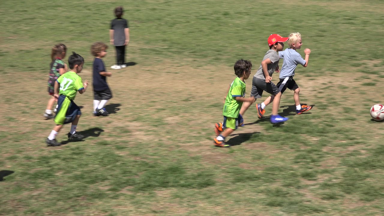 Young boys run across a patchy grass field in San Jose del Cabo, Mexico, with a soccer ball ahead of them and another child standing in the background.
