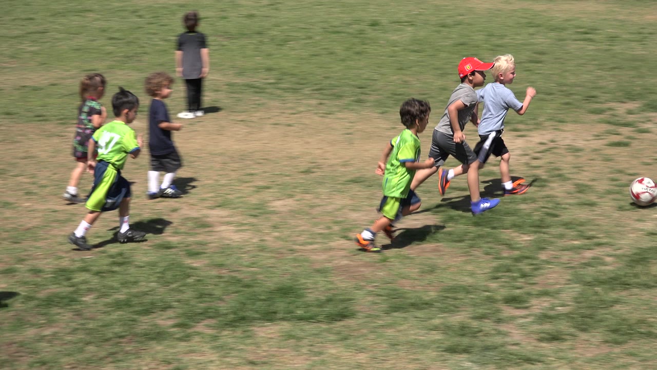 Several boys continue running toward a white and red soccer ball on a sunlit field in San Jose del Cabo, Mexico, with patchy grass filling the frame.