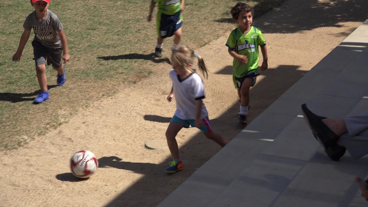 Children running near a soccer ball on a dirt path and grass.
