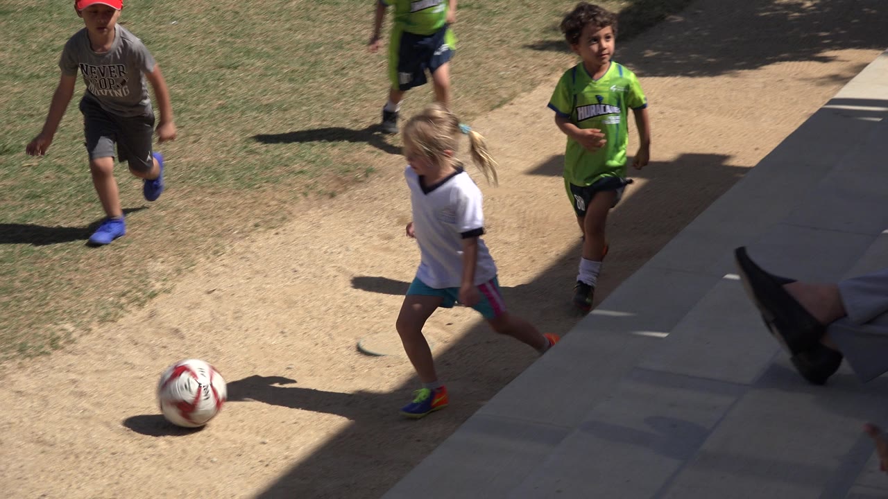 Children in San Jose del Cabo, Mexico move toward a white soccer ball near the edge of shade, with a girl in a white shirt crossing the path and boys behind her.