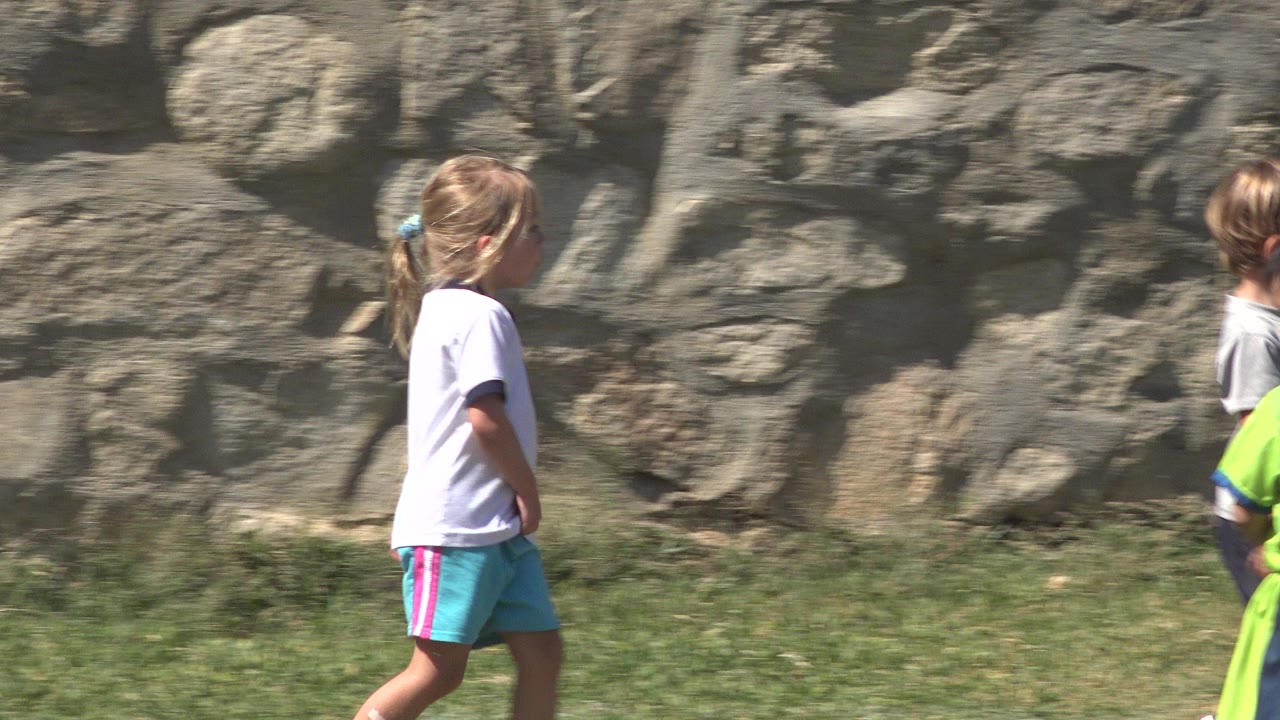 Blonde child walking across grass beside a stone wall.