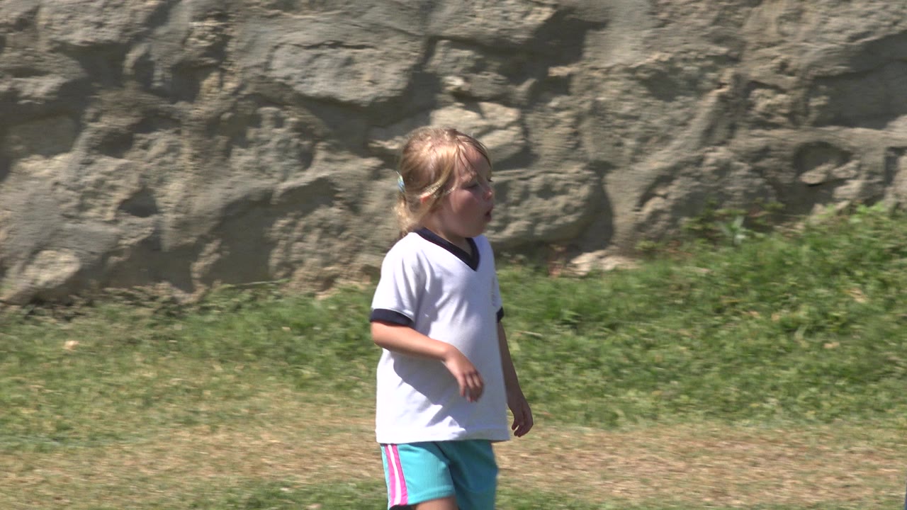 A young girl in a white shirt and turquoise shorts walks across a grassy field in San Jose Del Cabo, Mexico, with a rough stone wall behind her.