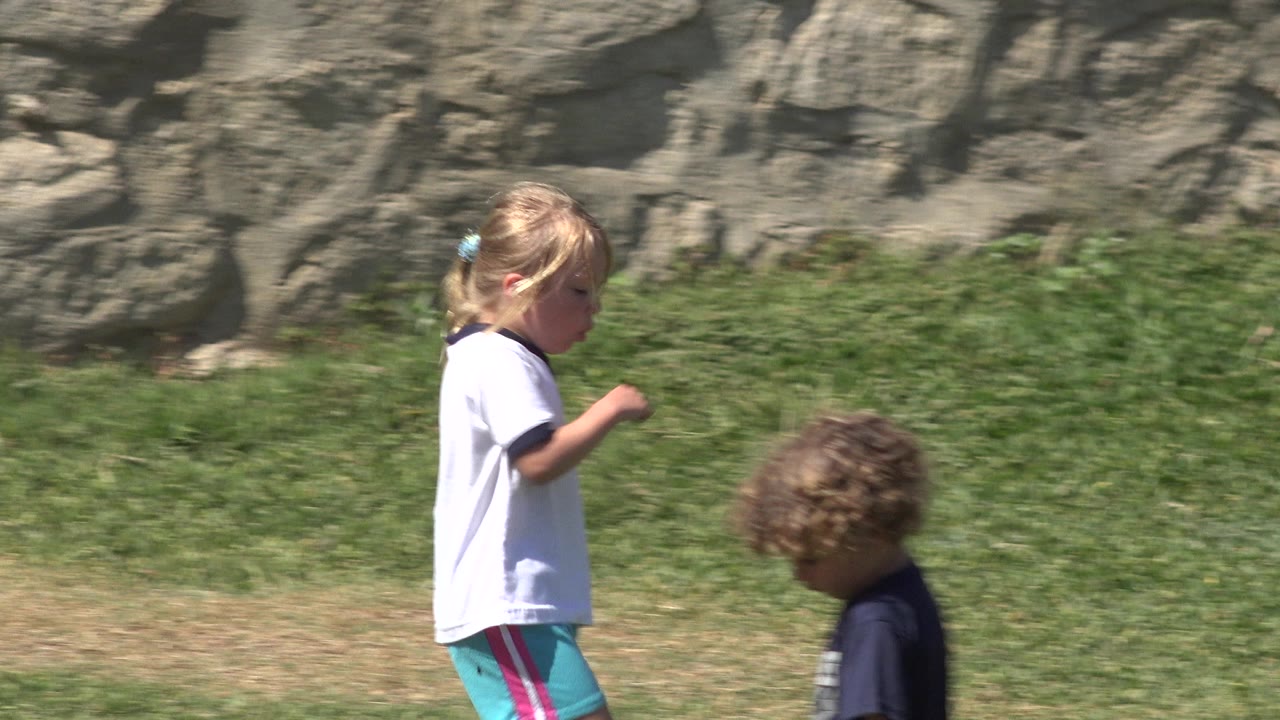 Young girl walking past another child on a grassy field.