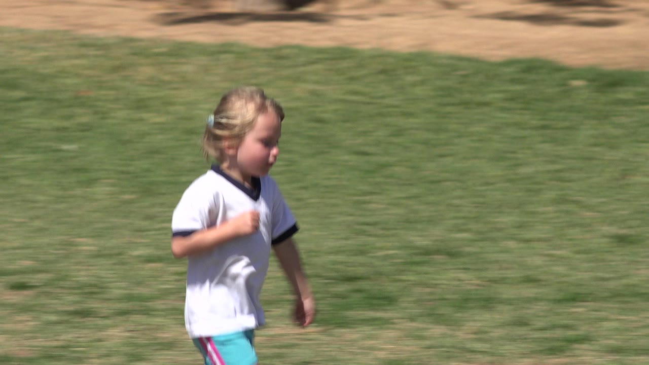 The girl continues running through the center of the frame on the green lawn, with her white shirt bright against the darker grass.