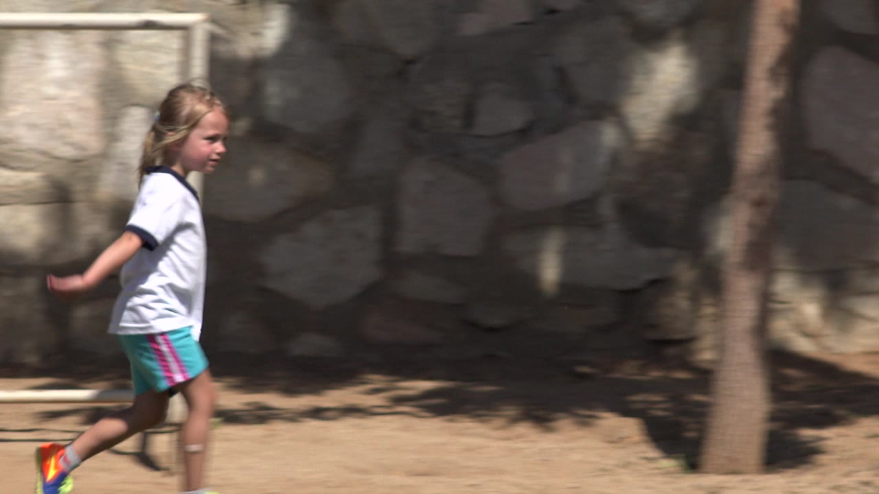 A young girl in a white shirt and turquoise shorts walks left to right in San Jose del Cabo, with a stone wall and tree shadows behind her.