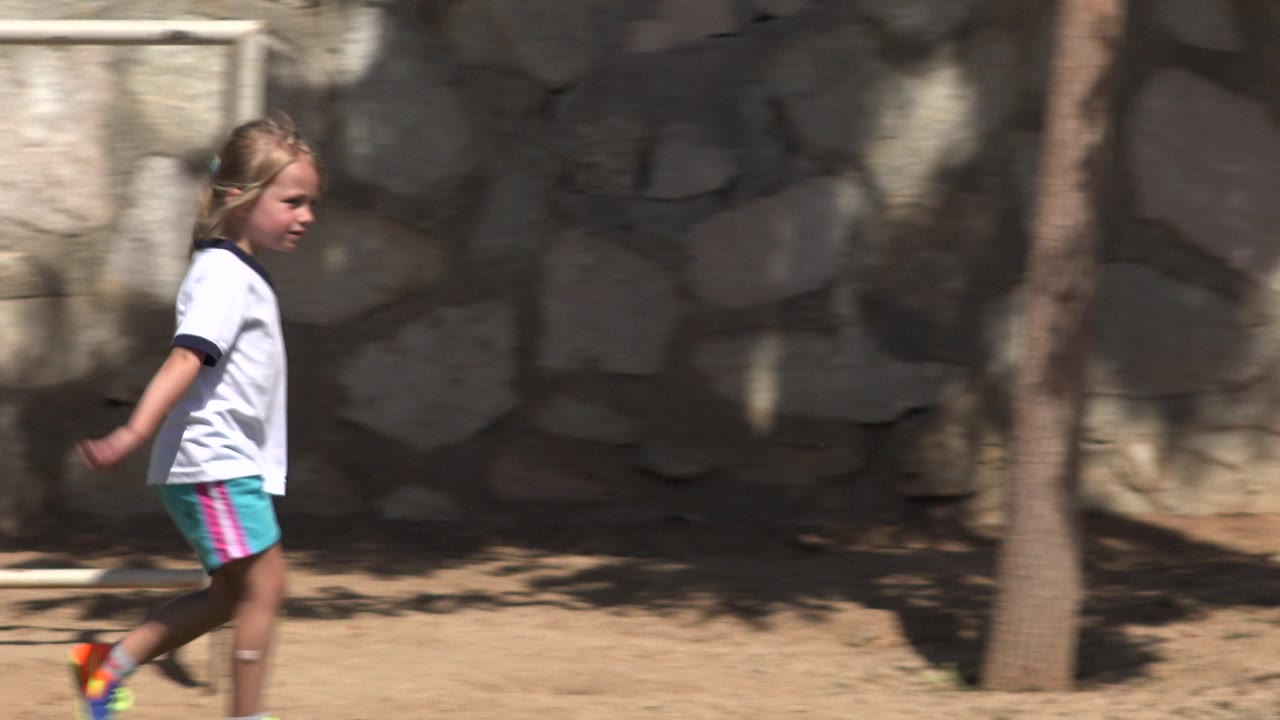The girl continues along the curb in San Jose del Cabo, passing a narrow tree trunk with the stone wall filling the background.