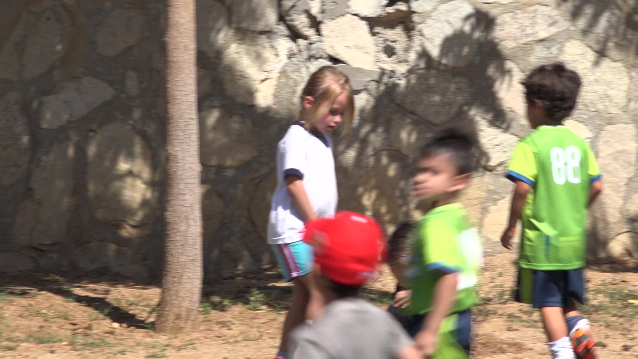 In San Jose del Cabo, Mexico, the girl looks down near the grassy edge as a boy in a green jersey with a visible number faces away by the stone wall.