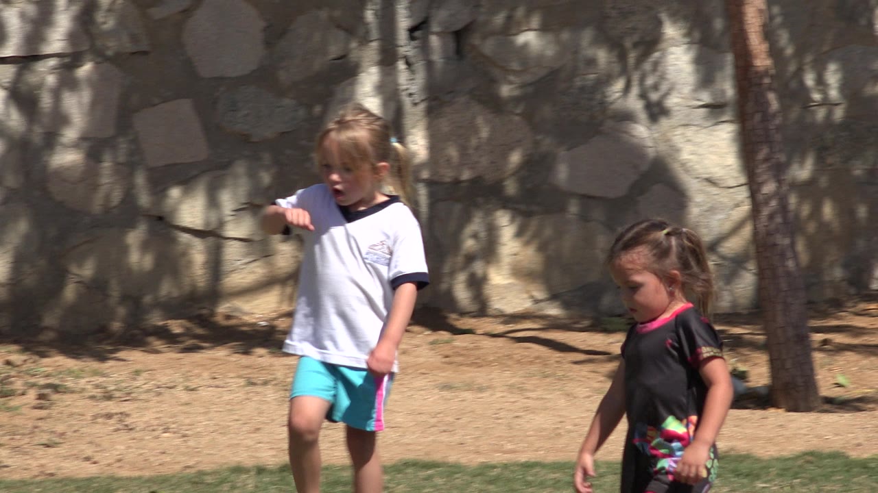 Two young girls continue across the grass in San Jose Del Cabo, Mexico, while the child in the white shirt points ahead near the stone wall.