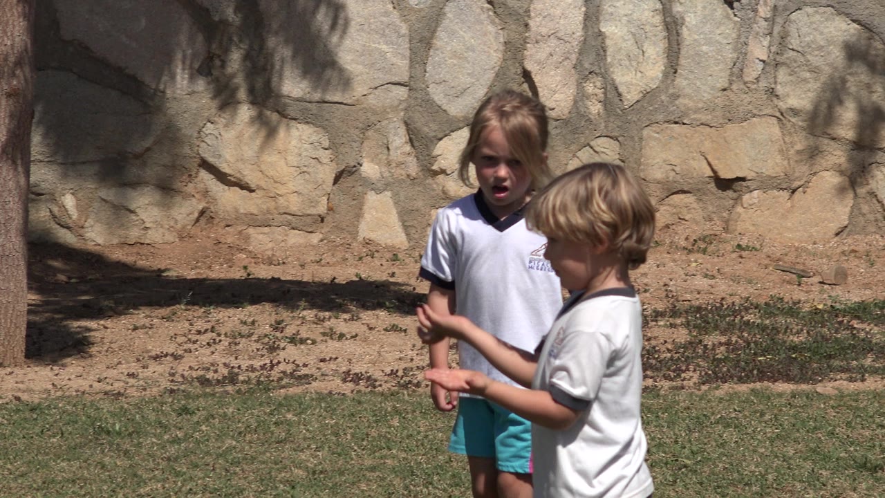 At 0:01 in San Jose del Cabo, Mexico, the children are still near each other on the grass, with the girl facing forward and the boy turned sideways against the same stone wall background.