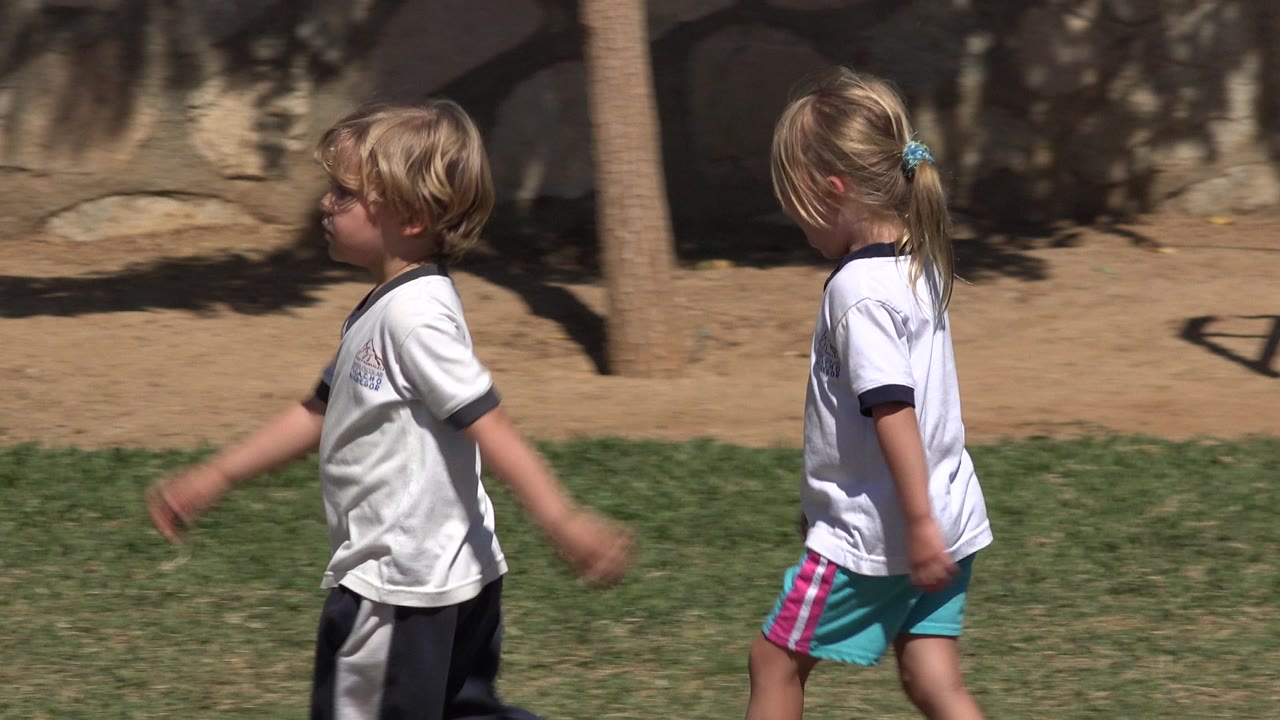 At the start of the clip, two children walk through a sunlit park in San Jose del Cabo, with grass below them and a stone wall behind.
