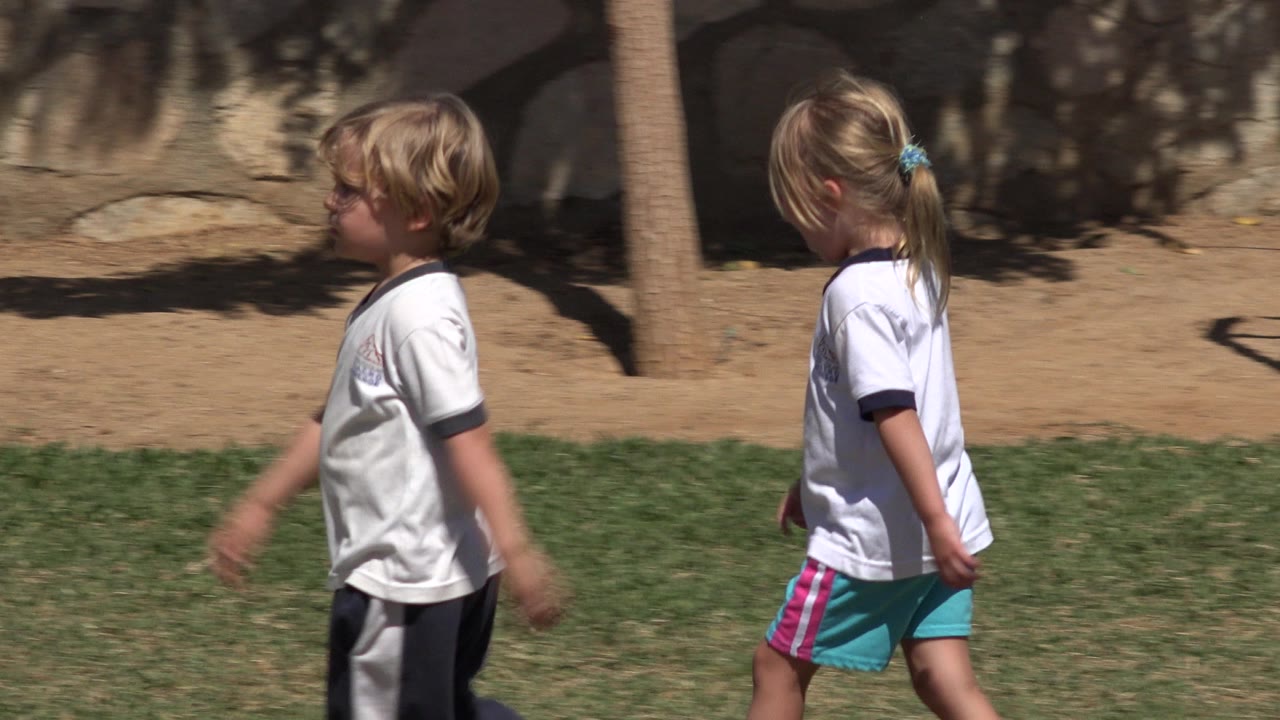 A girl with a ponytail walks rightward in San Jose del Cabo, wearing a white shirt and turquoise shorts near grass, bare soil, and a stone wall.