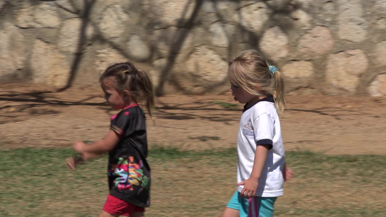 The girls continue across the grass in San Jose del Cabo, Mexico, with the darker-haired child slightly ahead and the blonde child close behind.