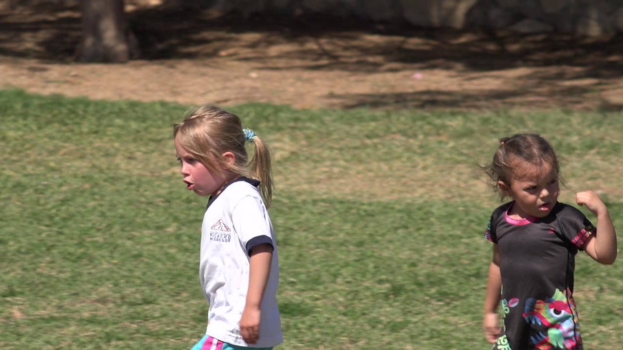 The girls continue walking across the green lawn in San Jose del Cabo, Mexico, one looking forward and the other lifting an arm near her shoulder.