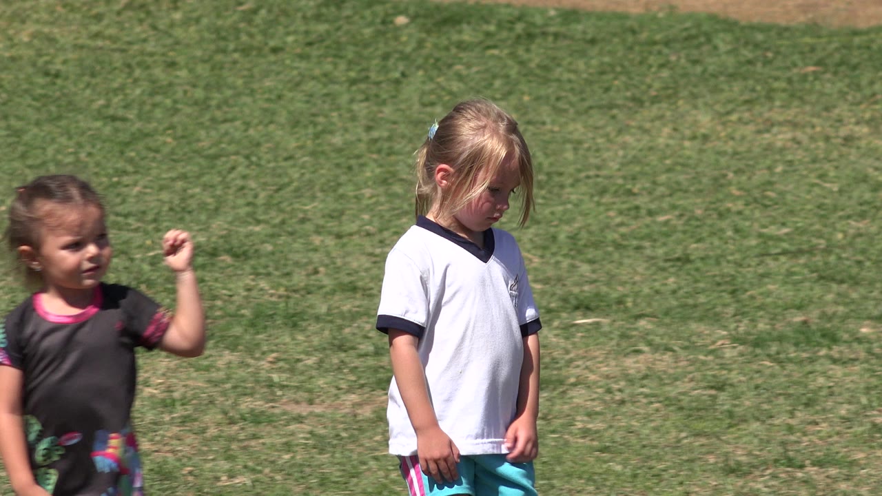 A young child in a dark shirt raises a hand on the grass field while a blond child in a white shirt looks downward in San Jose del Cabo, Mexico.