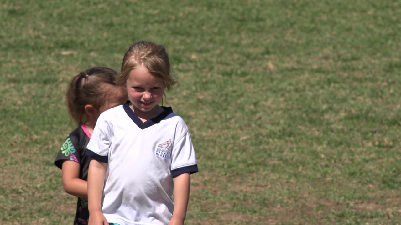 Two young girls stand close together on a grass field in San Jose del Cabo, Mexico, with the girl in a white jersey facing the camera and another child partly behind her.