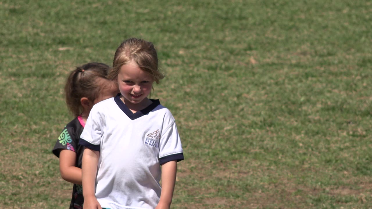 A young girl in a white sports jersey stands in front on the grass field in San Jose del Cabo, Mexico, while a second child is partially hidden behind her shoulder.