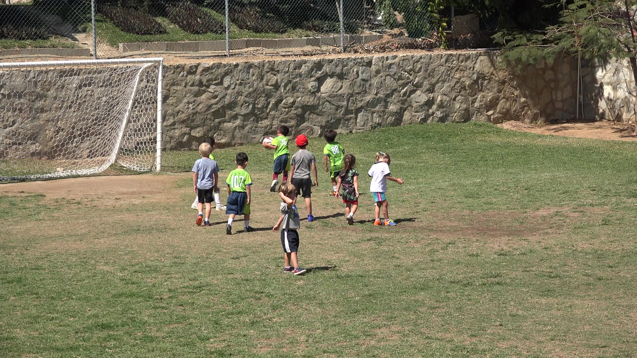 A small group of children in bright shirts stands near the left-side soccer goal on the field in San Jose del Cabo, Mexico, while another child is lower in the frame.