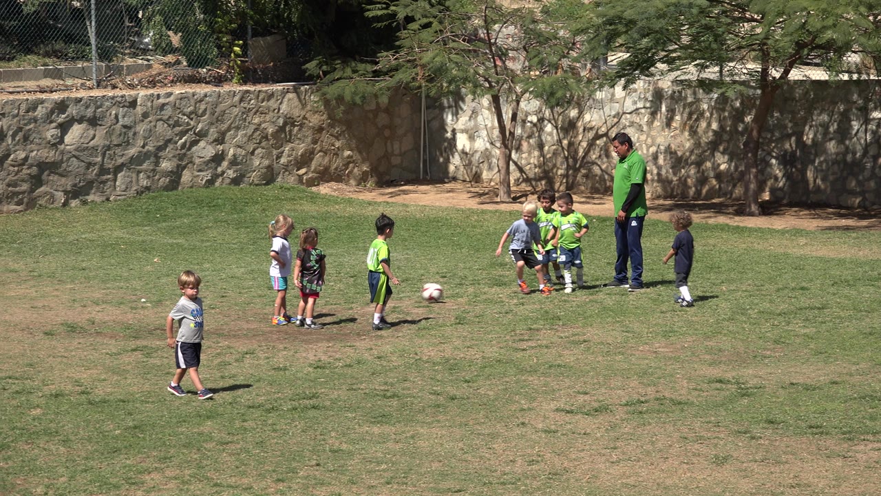 Several very young children cluster near the soccer ball in San Jose del Cabo, Mexico, while an adult stands to the right and other children wait across the grass.