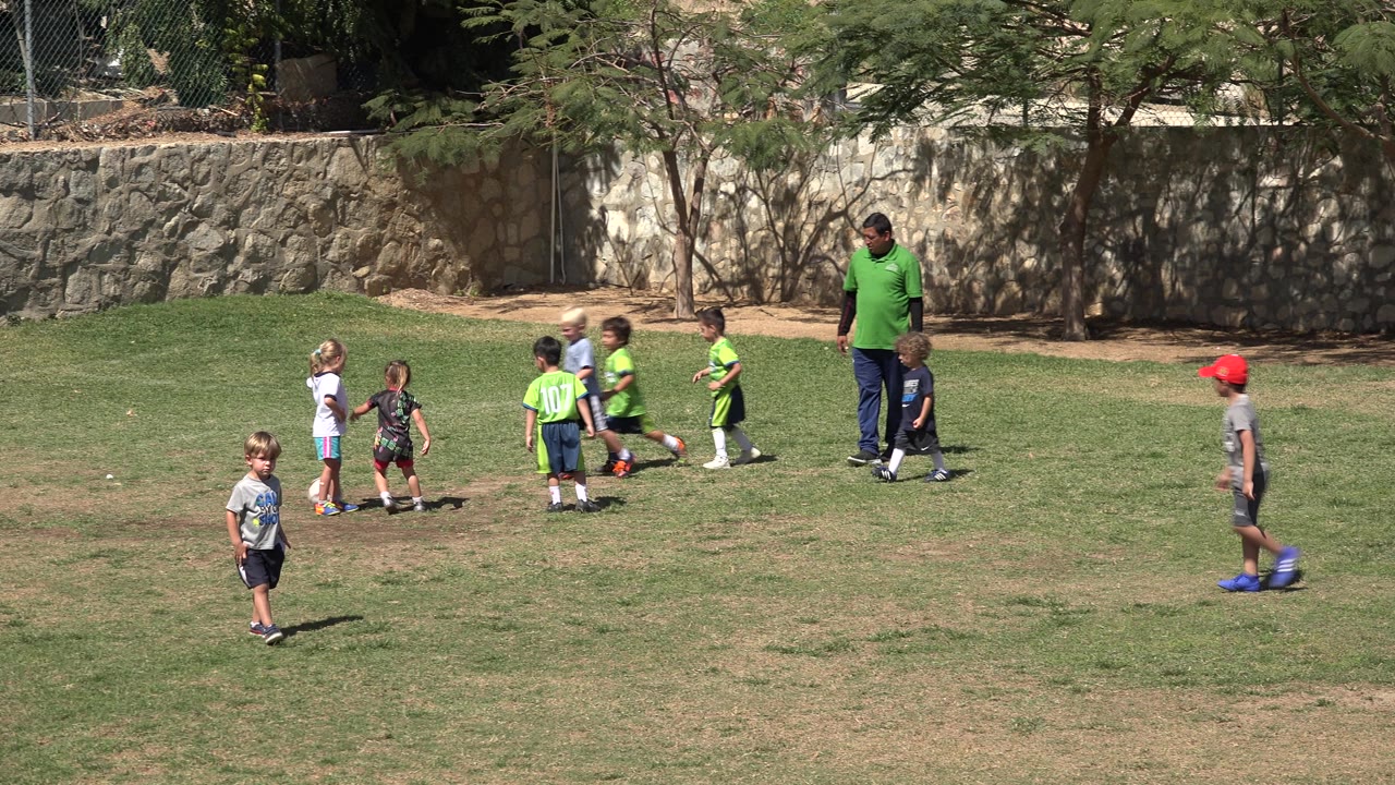 Children in soccer clothes move across a lawn with an adult nearby.