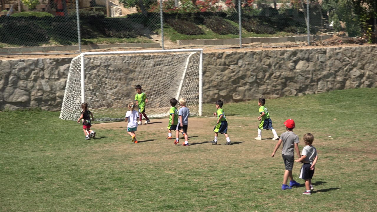 Children in green and white shirts cluster by the goal in San Jose del Cabo, Mexico, while two boys stand on the grass in the foreground.