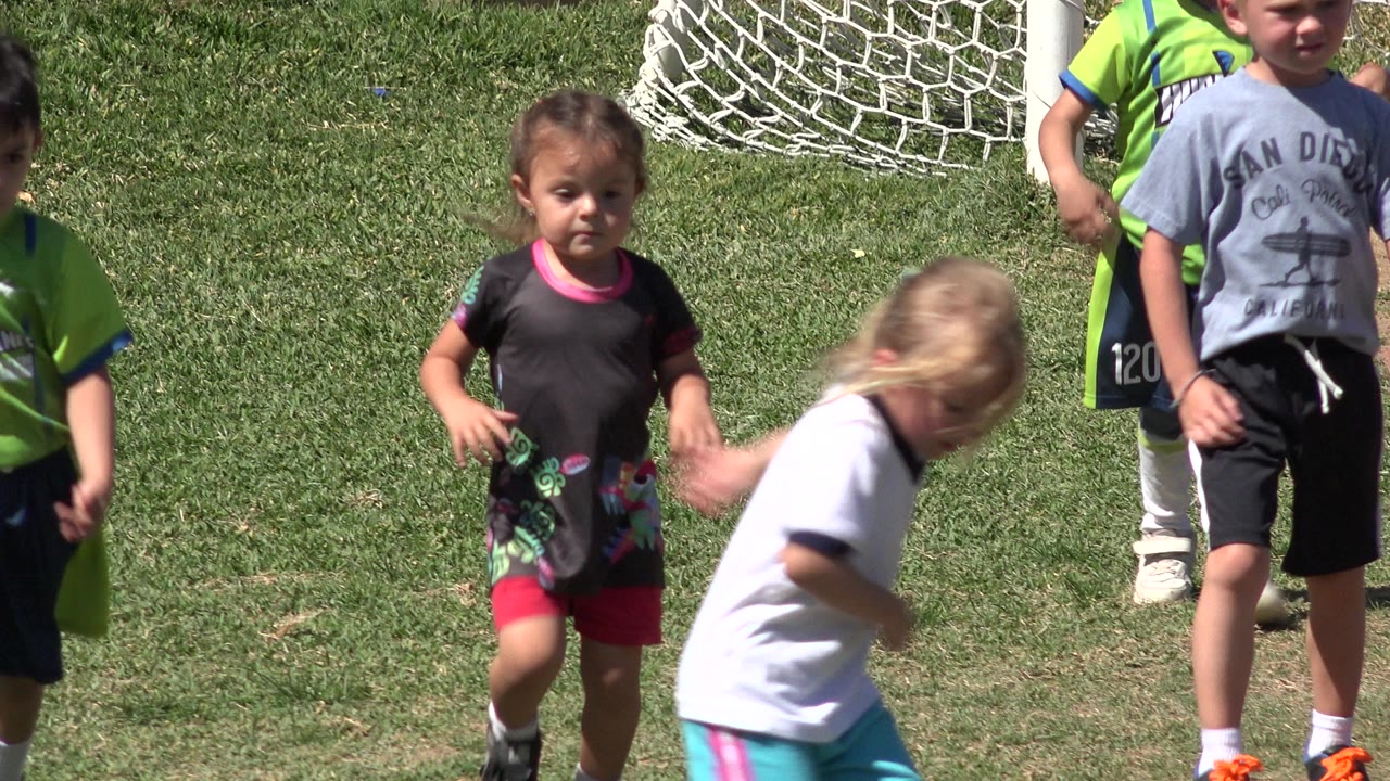 Children run on a grassy soccer field in San Jose del Cabo, Mexico, with a girl in black and pink near the foreground and a white goal net behind them.