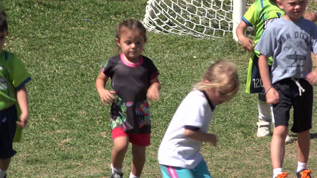 A young girl in a dark shirt runs toward camera while other children in soccer clothing move around her on the grass in San Jose del Cabo, Mexico.