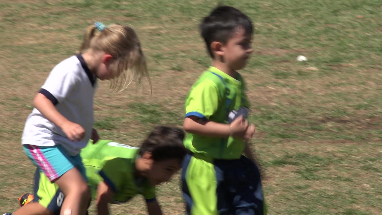 A blonde child in a white shirt and a child in a green jersey move near another child bent low on the grass field in San Jose del Cabo, Mexico.