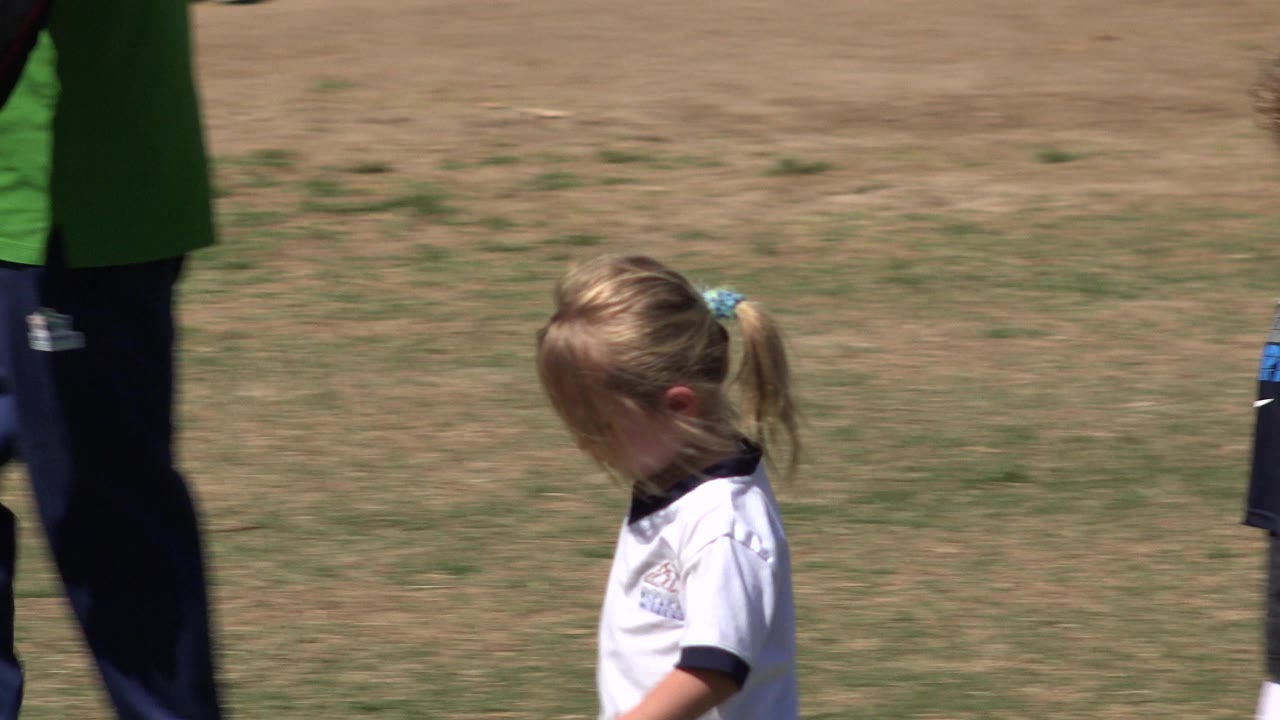 A young blonde girl in a white shirt walks across a sunlit grass field in San Jose del Cabo, Mexico, with part of an adult in green visible at the left edge.