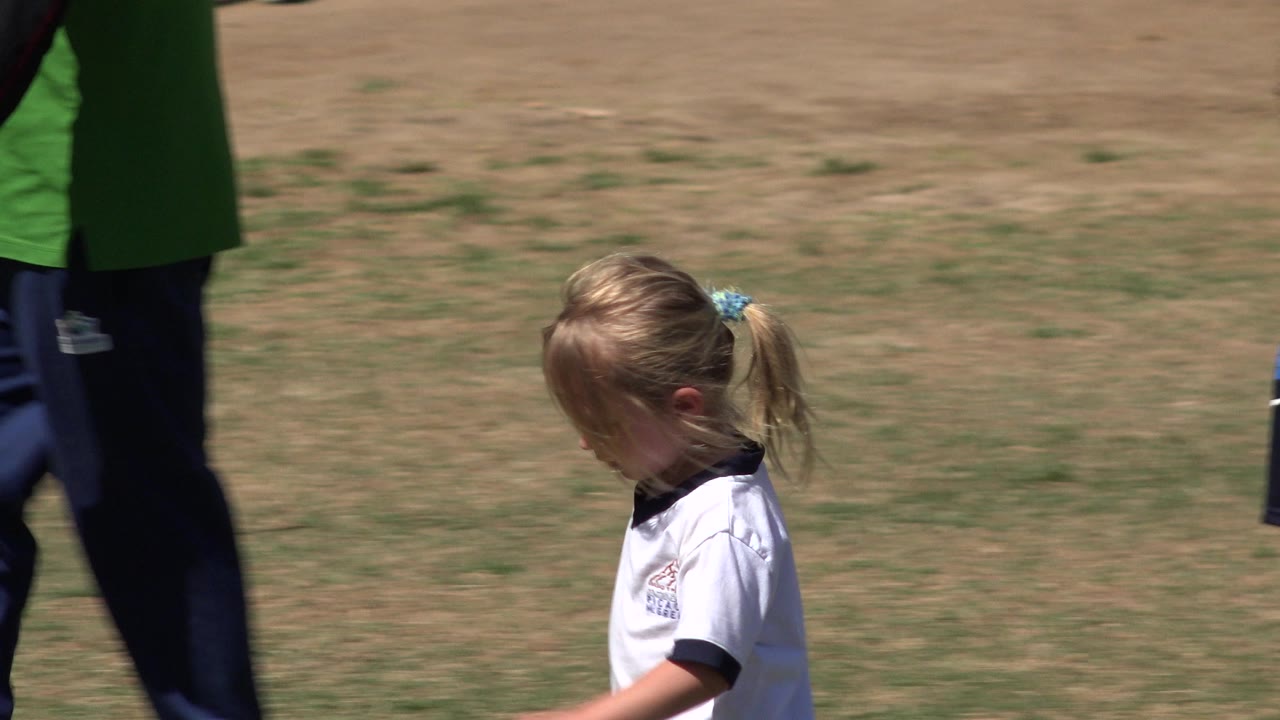 The girl looks downward as she moves across the grassy field in San Jose del Cabo, Mexico, while a cropped adult remains behind her.