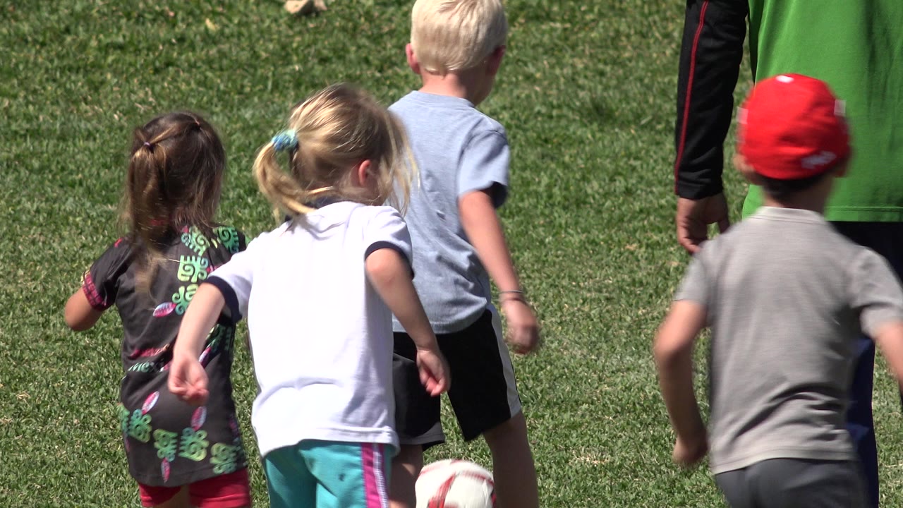 Children in casual clothes chase a red and white soccer ball across a sunny grass field in San Jose del Cabo, Mexico, with an adult in a green shirt partly visible ahead.