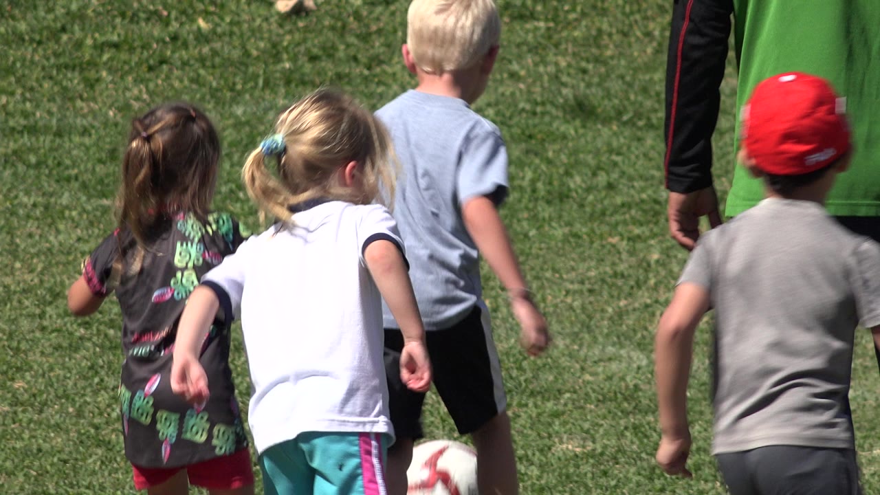 A small group of children runs from behind toward the soccer ball on a grass field in San Jose del Cabo, Mexico, with a child in a red cap near the center.
