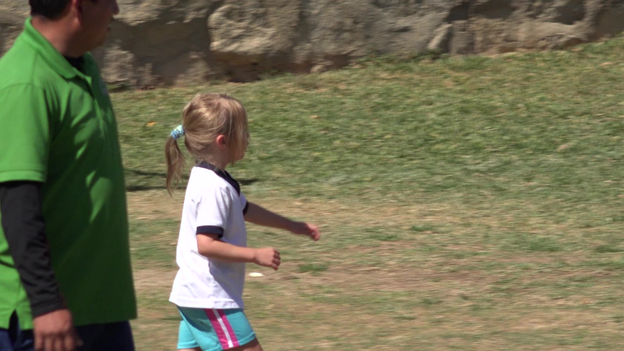 Young girl running on grass beside an adult in a green shirt.
