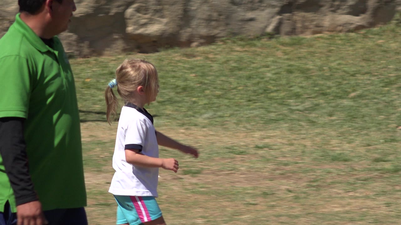 At 0:01 in San Jose Del Cabo, Mexico, the girl runs farther right across the grass as the adult in the green polo remains partially cropped in the foreground.