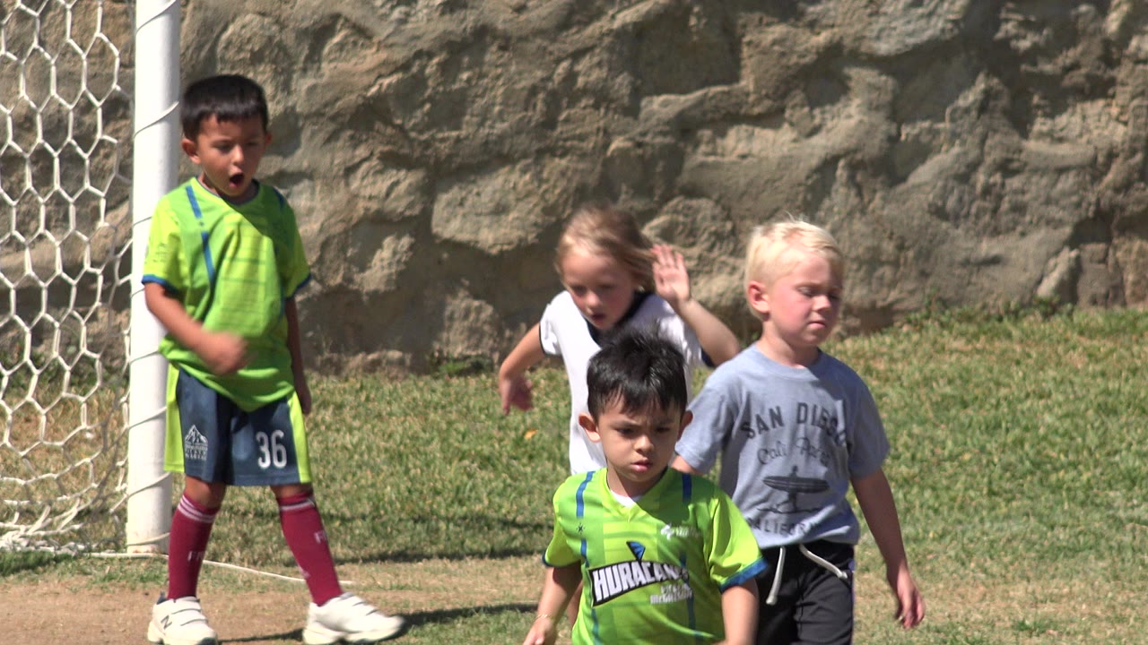 Children stand and move near a white soccer goal in San Jose Del Cabo, Mexico, with a blonde child in a gray shirt and other players in bright green jerseys visible on the grass.