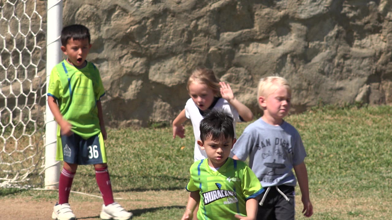 A group of children in San Jose Del Cabo, Mexico gathers around the goal area, including a child in a white jersey raising an arm and a blonde child in gray walking nearby.