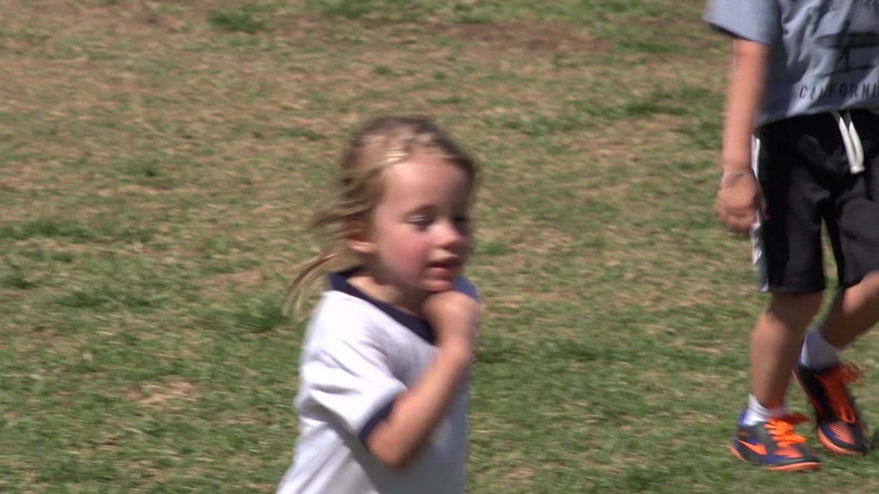 A young blonde girl in a white jersey runs across a sunlit grass field in San Jose del Cabo, Mexico, while another child is cropped at the right edge.