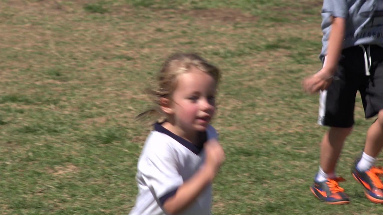 The girl jogs in a white jersey on the San Jose del Cabo field as another child in gray and black sportswear passes partly cropped in the background.