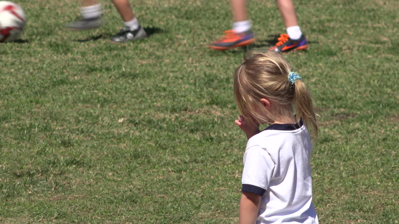 Blonde child in a white soccer shirt standing on a grass field.