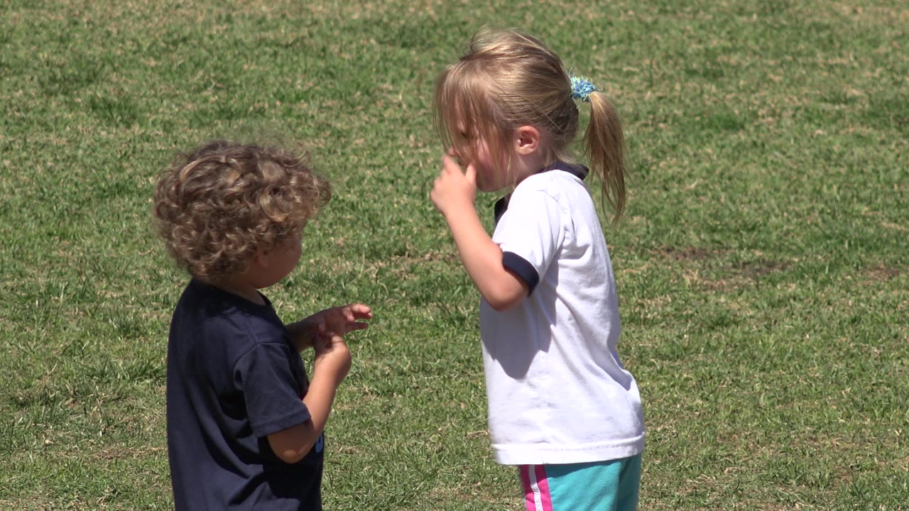 Two young children face each other on a sunlit grassy field in San Jose del Cabo, Mexico, with the girl in a white shirt turned in profile.