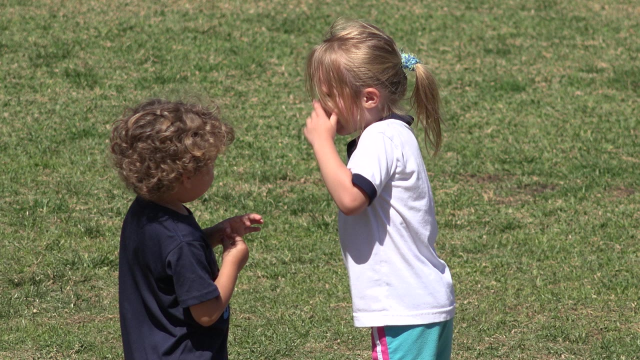 The curly-haired child stands with his back partly to camera while the girl in a white shirt raises a hand near her face on the grassy field in San Jose del Cabo, Mexico.