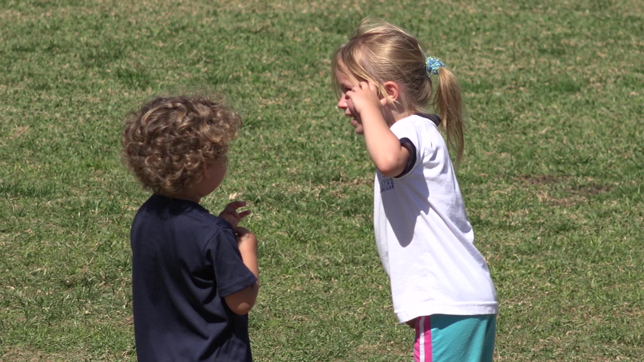 The girl in the white shirt touches her face as the curly-haired child looks toward her on the open grass in San Jose del Cabo, Mexico.