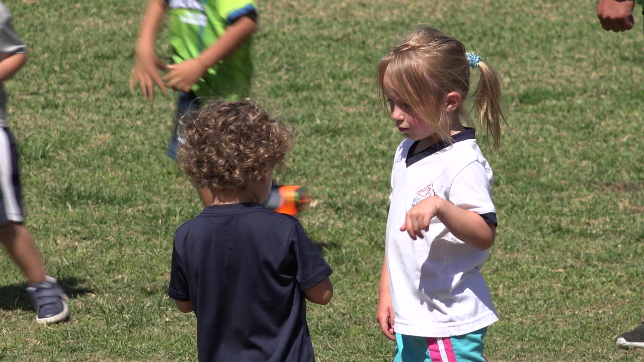 At 0:01, the blonde girl looks toward the curly-haired child while cropped children in green and gray clothing move along the edges of the San Jose del Cabo grass field.