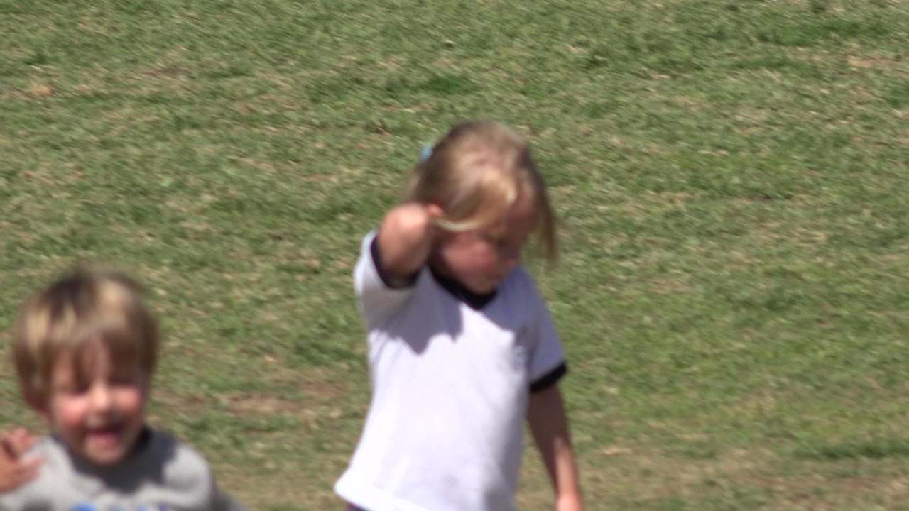 The young girl in a white jersey moves closer to camera on the grass field, her face partly lowered and softened by motion blur.