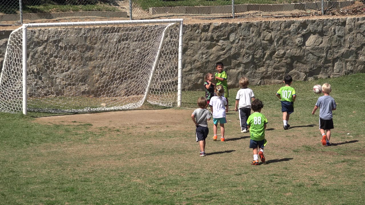 Several young children stand and move near a white soccer goal on a sunlit field in San Jose del Cabo, Mexico, with a rock wall behind them.
