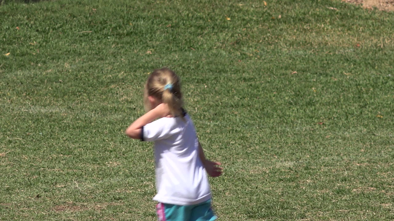 A blonde child in a white shirt and teal shorts runs across a grass field in San Jose del Cabo, Mexico, with no goal visible in this opening frame.