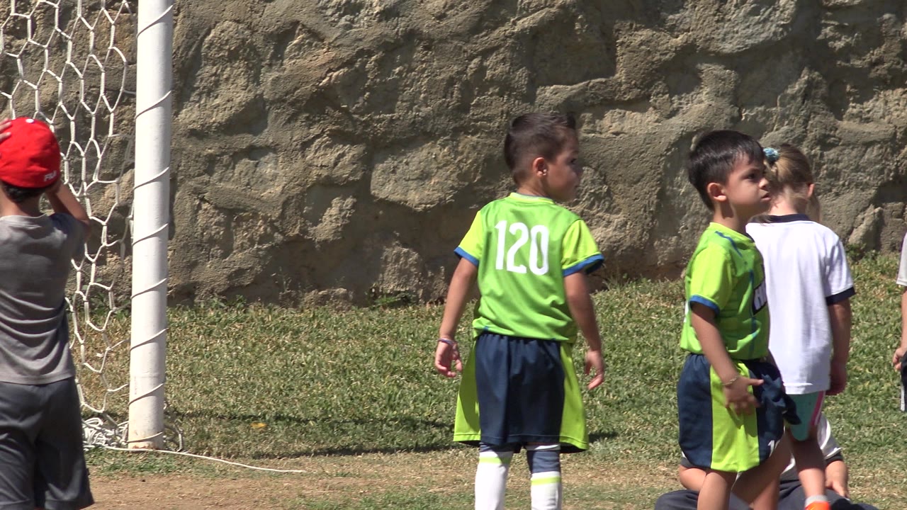 Young players in green uniforms and a child in a white shirt gather near the goalpost on the San Jose Del Cabo grass field.
